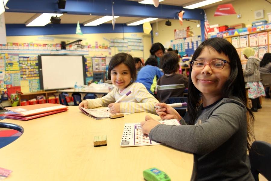 Two students smile for a photo while sitting at a desk in a classroom